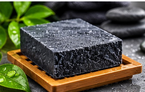 Charcoal soap bar on wooden dish with water droplets and green leaves
